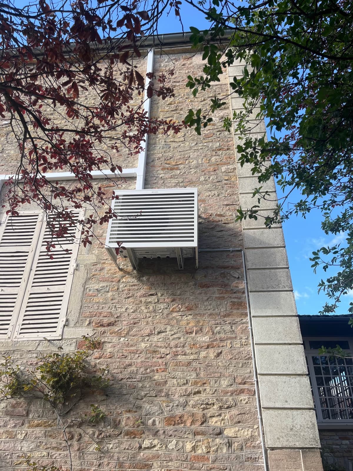 Groupe extérieur de climatisation habillé d'un cache-clim à lames sur la façade en pierre d'une demeure à Prissé