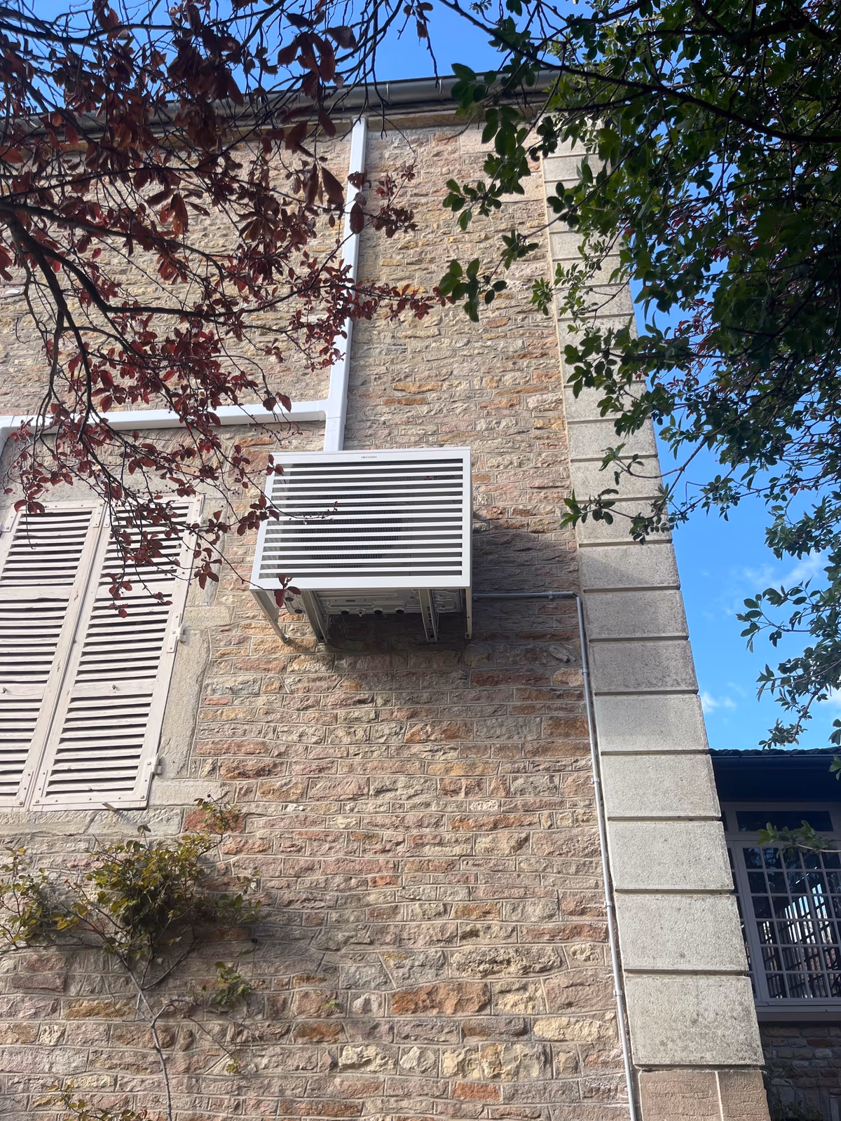 Groupe extérieur de climatisation habillé d'un cache-clim à lames sur la façade en pierre d'une demeure à Prissé